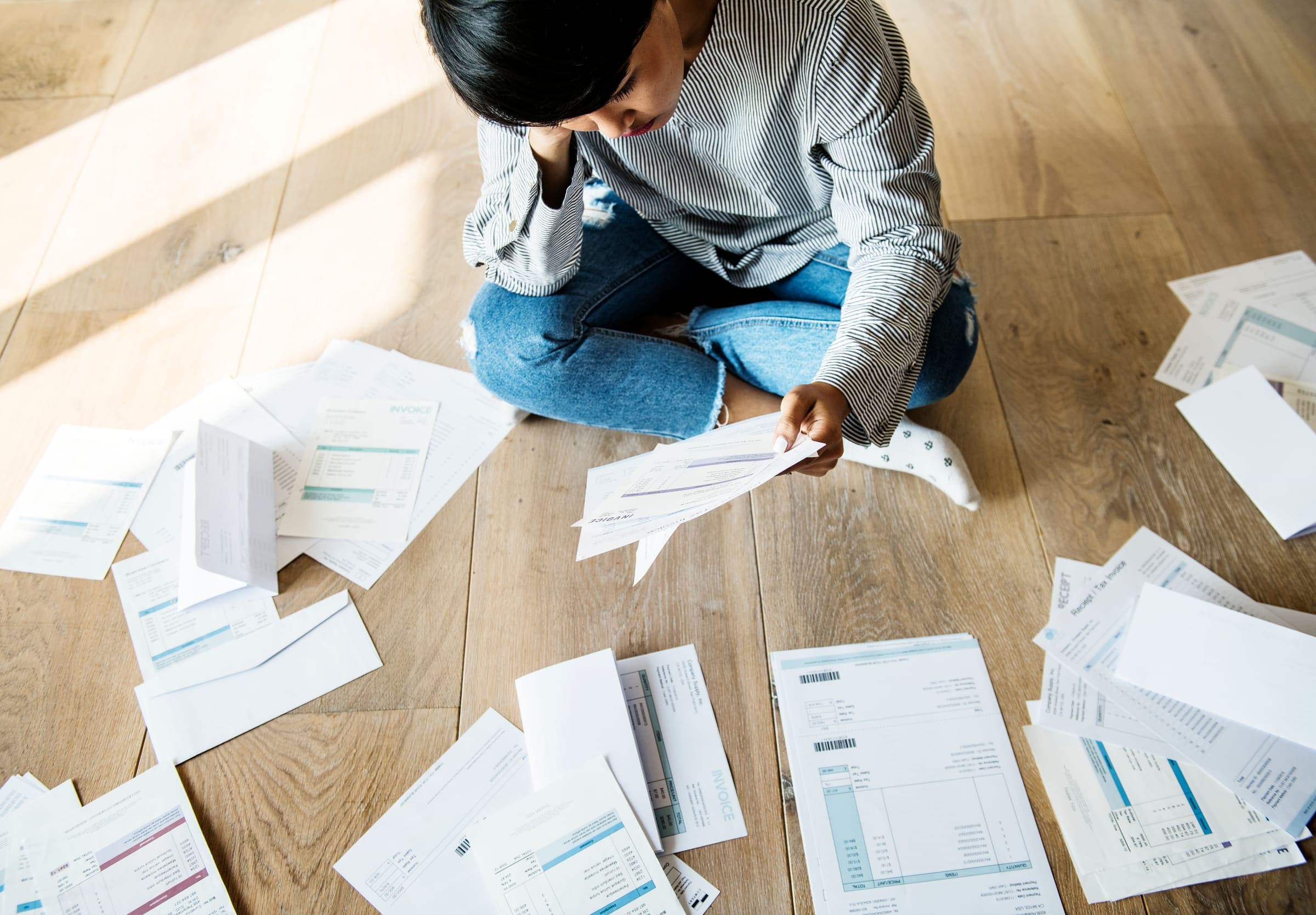A person sitting on a wooden floor surrounded by opened letters and bills, reading one of them