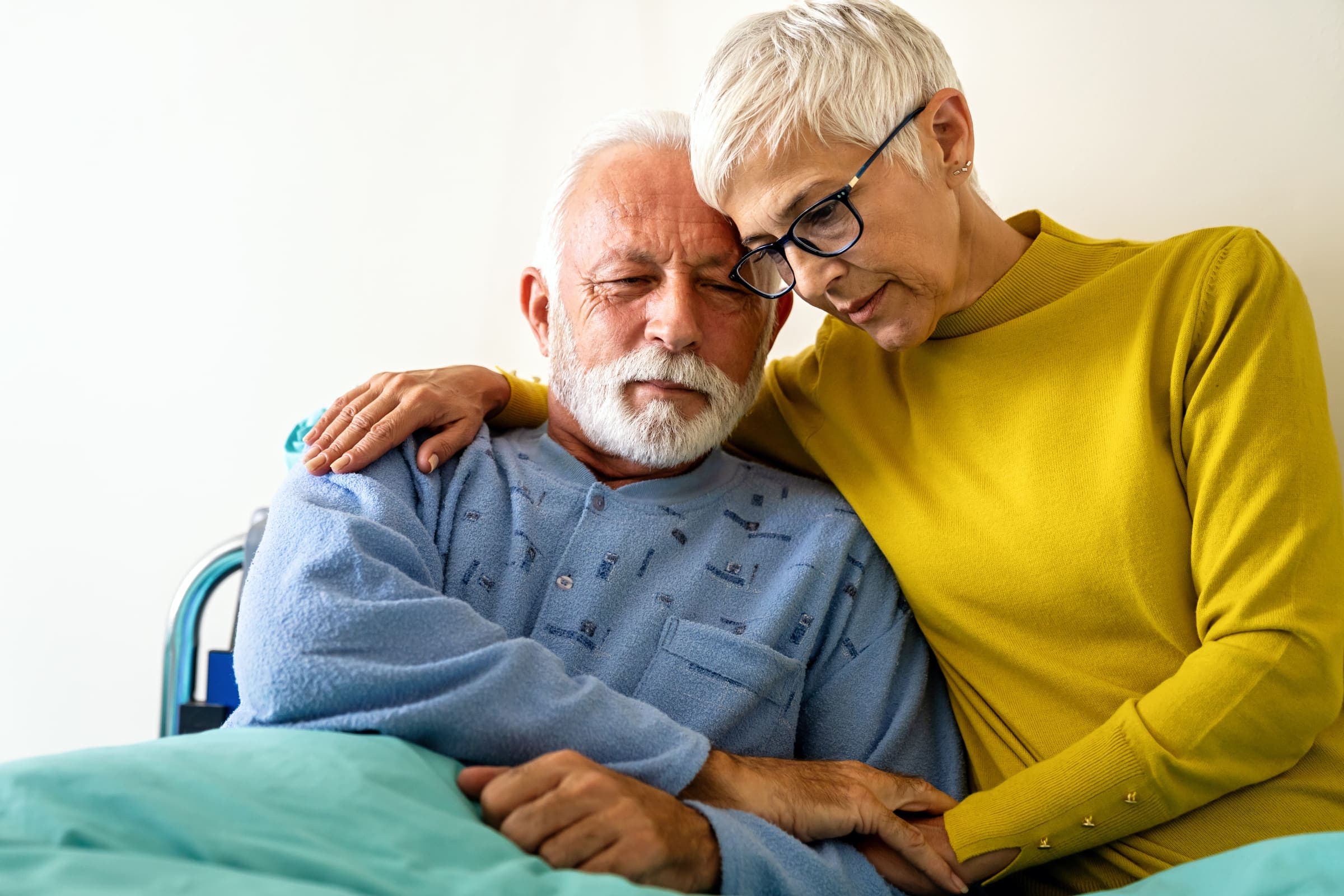 An older man in a wheelchair resting his head against a woman who has an arm around his shoulder
